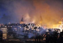 A large crowd of police officers in helmets facing a smoky protest scene at night