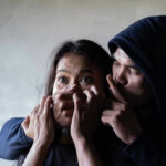 Armed Bandits SLAUGHTER Staff — 25 Girls VANISH Man holding womans mouth, gesturing silence.