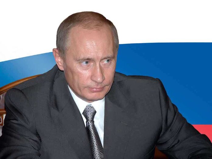 A serious-looking man in a suit sitting at a table with a Russian flag in the background
