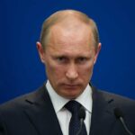 A serious-looking man in a suit at a press conference with a blue background