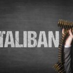 A hand holding a belt of bullets in front of the word TALIBAN on a dark background