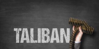 A hand holding a belt of bullets in front of the word TALIBAN on a dark background