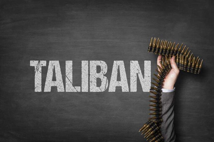 A hand holding a belt of bullets in front of the word TALIBAN on a dark background