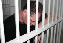 A man sitting in a jail cell with his head down, resting on the bars