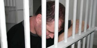 A man sitting in a jail cell with his head down, resting on the bars