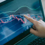 Hand pointing at stock market graph on laptop.