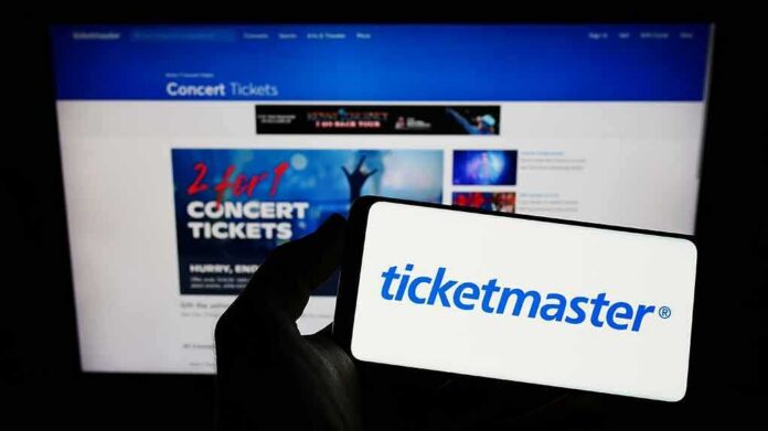 A hand holding a smartphone displaying the Ticketmaster logo in front of a computer screen showing concert ticket promotions