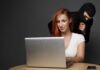 A woman using a laptop with a shadowy figure behind her, suggesting a cybersecurity threat