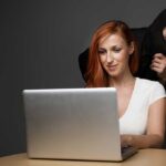 A woman using a laptop with a shadowy figure behind her, suggesting a cybersecurity threat