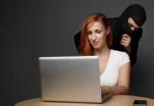 A woman using a laptop with a shadowy figure behind her, suggesting a cybersecurity threat