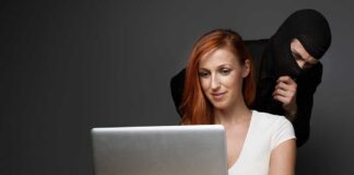 A woman using a laptop with a shadowy figure behind her, suggesting a cybersecurity threat
