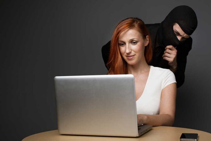 Man,In,A,Balaclava,Furtively,Watching,An,Unsuspecting,Female,Office A woman using a laptop with a shadowy figure behind her, suggesting a cybersecurity threat
