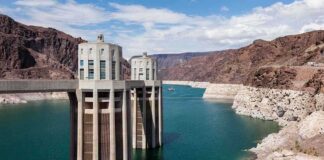 A large dam structure with two towers over a blue water reservoir surrounded by rocky mountains