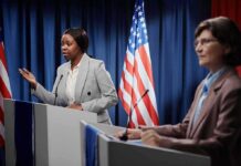 Two women at podiums during a debate with U.S. flags behind them